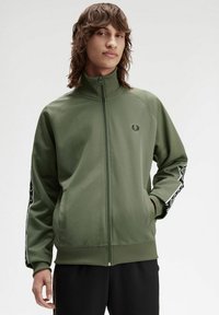 Giacca zip-up verde oliva con colletto alto, polsini a costine e due tasche laterali; presenta un logo nero e strisce laterali bianche a contrasto.