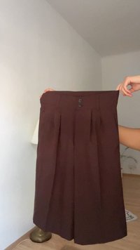 Jupe plissée marron foncé avec deux boutons à la taille, tenue par deux mains dans une pièce avec un sol en bois et des murs blancs.