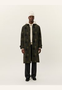 Lungo cappotto di lana in un motivo a quadri verde scuro e nero, con colletto a scollo e bottoni, indossato sopra un hoodie beige e pantaloni scuri.