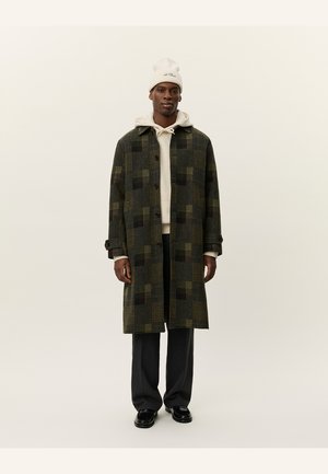 Abrigo largo de lana en un patrón de cuadros verde oscuro y negro, con cuello de muesca y botones, usado sobre una sudadera beige y pantalones oscuros.