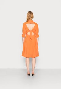 Vestido laranja com mangas curtas bufantes, costas recortadas com um detalhe de laço e uma saia plissada com um padrão texturizado.