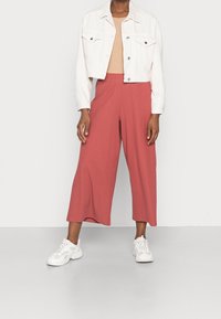 Giacca di jeans bianca tagliata sopra una maglietta a coste beige, abbinata a pantaloni a gamba larga color ruggine e sneakers bianche.