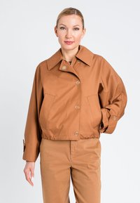 Giacca corta marrone con ampio colletto, chiusura frontale con bottoni e tasche laterali, realizzata in tessuto liscio. Indossata con pantaloni dello stesso colore.