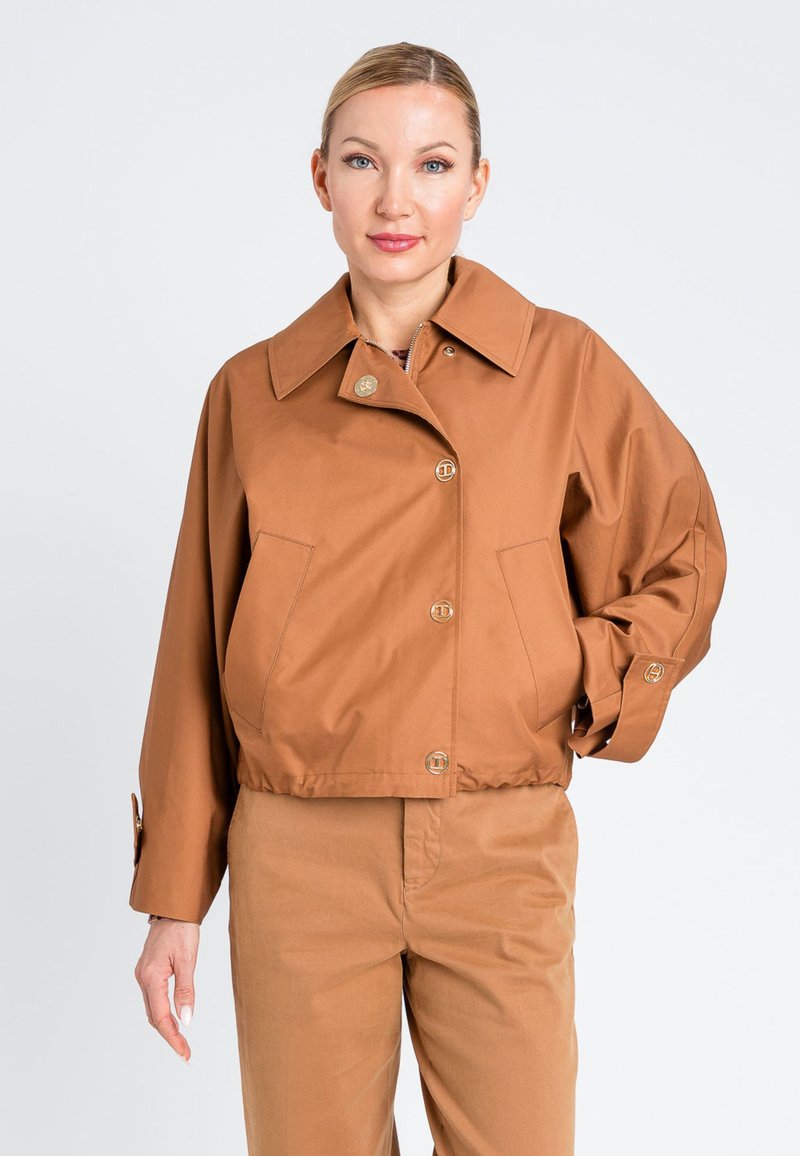 Giacca corta marrone con ampio colletto, chiusura frontale con bottoni e tasche laterali, realizzata in tessuto liscio. Indossata con pantaloni dello stesso colore.