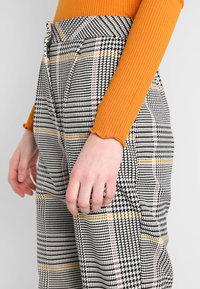 Hand an der Hüfte, trägt schwarze und weiße Hahnentritt-Hosen mit gelben und rosa Streifen, kombiniert mit einem orangen, gerippten Langarmoberteil.