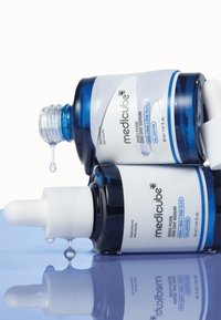 Dwie szklane buteleczki z serum z niebieskimi i białymi etykietami, zakraplaczami oraz kroplami cieczy spływającymi na powierzchnię odbijającą. Opakowanie zawiera tekst i branding.