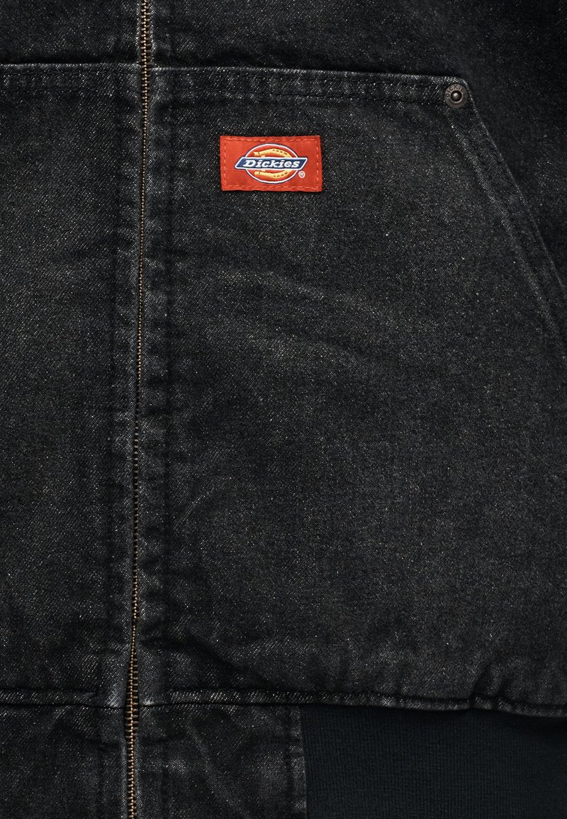 Zwarte spijkerjas met ritssluiting, zichtbare stiksel details en een oranje stoffen label met het Dickies-logo op de bovenste linkerzijde.