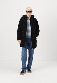 Manteau doudoune noir avec capuche, coutures verticales et poches latérales, associé à un pull rayé et un pantalon à motifs bleus. Chaussures : baskets noires.