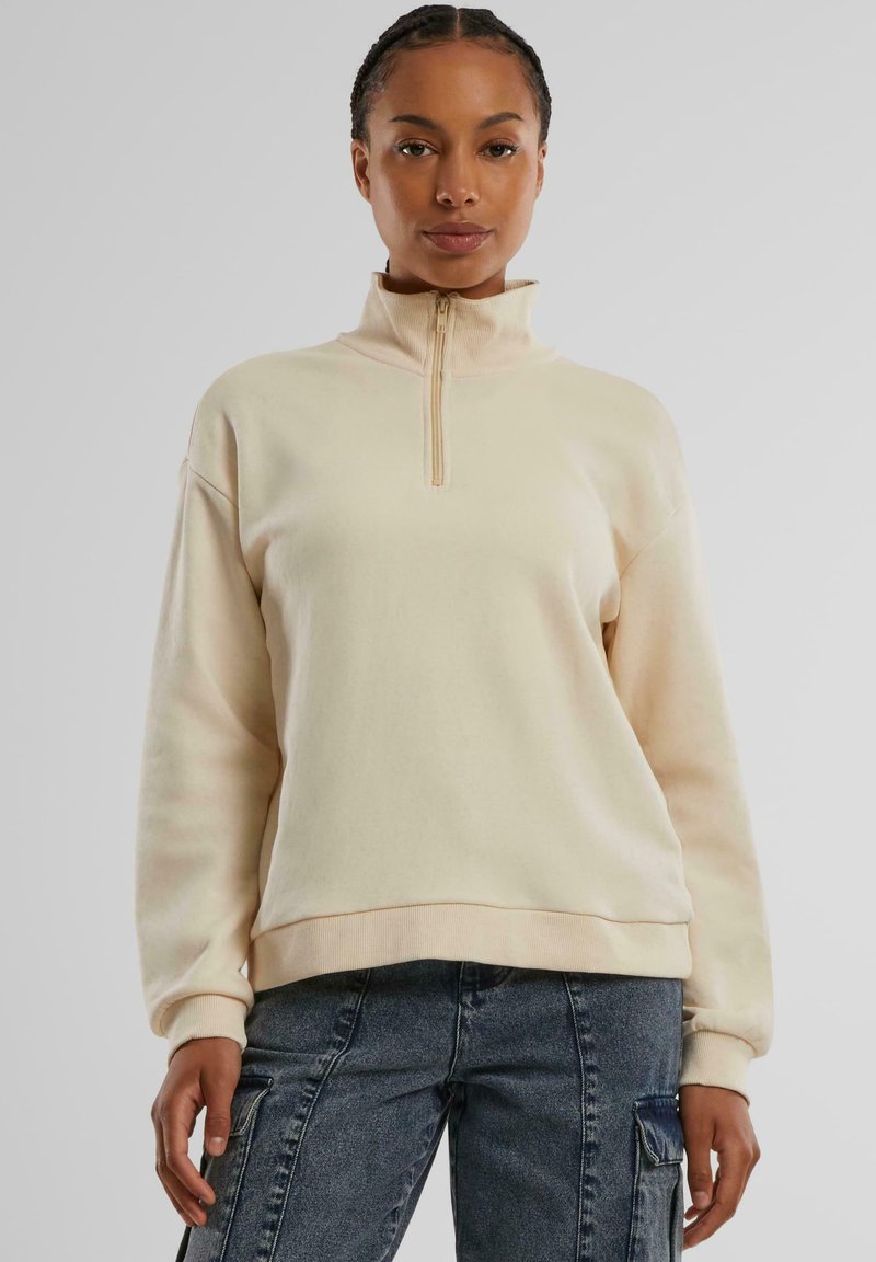Sweatshirt couleur crème avec un col montant et une fermeture éclair demi-longueur, présentant une coupe décontractée et des poignets côtelés. Tissu doux avec une texture lisse.