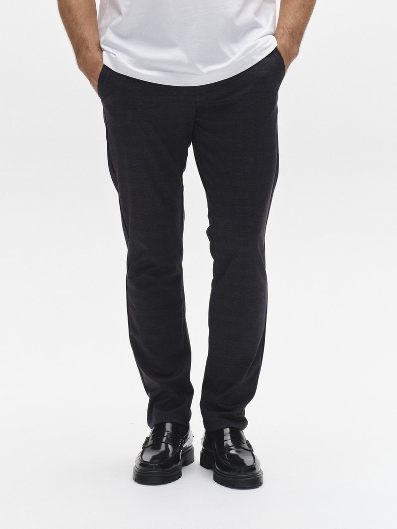 Sötétkék, texturált anyagból készült, testhez simuló, oldalanként zsebes nadrág, fekete, fényes cipővel és fehér pólóval viselve.