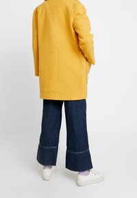 Cappotto giallo texture con vestibilità rilassata, abbinato a pantaloni a gamba larga in denim scuro e sneakers bianche. Dettagli a vista sull'orlo dei pantaloni.