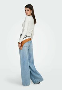 Haut blanc à manches longues, jean bleu clair à jambes larges, et sac ceinture rayé marron et noir. Tissu lisse et coupe décontractée.