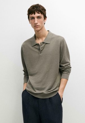 Jonge man met bruin haar, gekleed in een olijfgroene poloshirt met lange mouwen en donkerblauwe broek, staand met handen in de zakken tegen een witte achtergrond.