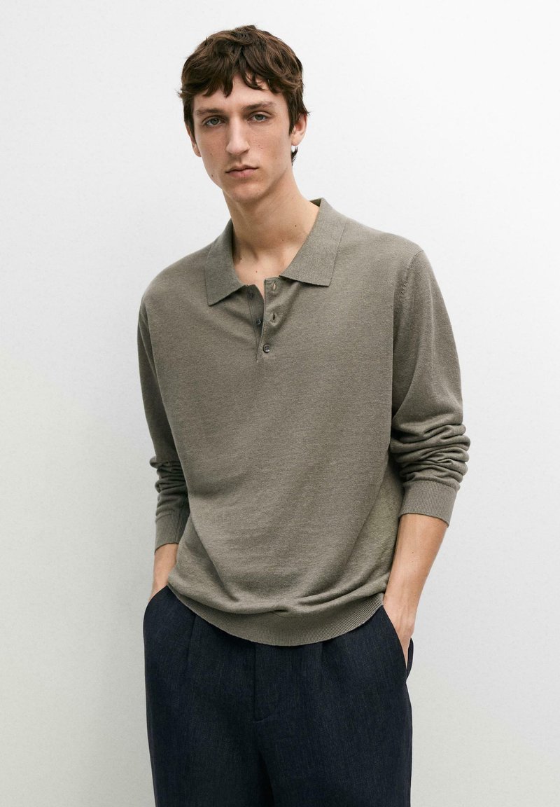 Jeune homme aux cheveux bruns portant un polo manches longues vert olive et un pantalon bleu marine, debout les mains dans les poches sur fond blanc.