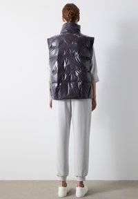 Gepolsterte Weste aus glänzendem dunkelgrauem Material mit hohem Kragen und ärmellosem Design. Kombiniert mit hellgrauen schmal zulaufenden Hosen und weißen Schuhen.