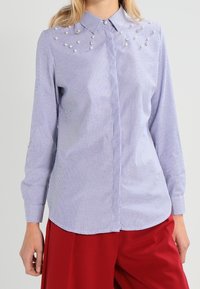 Striped button-up shirt in blue and white cotton, featuring pearl accents on shoulders and cuffs, long sleeves, and a rounded hem.