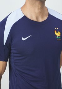 Námořnické modré sportovní tričko s světle modrými ramenními akcenty a strukturovanou tkaninou. Obsahuje logo Nike a vyšívaný emblém kohouta se hvězdami.