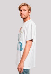 White cotton t-shirt with short sleeves featuring colorful text design: "CLUB" in blue, orange, and green. Model wears blue jeans.