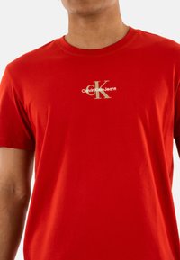 Camiseta de algodón roja con cuello redondo. Presenta un logo dorado de Calvin Klein Jeans en el pecho. Mangas cortas y una silueta ajustada.
