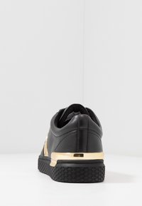 Zapatilla negra con suela texturizada y acento metálico dorado en el talón, vista desde atrás sobre un fondo blanco simple.