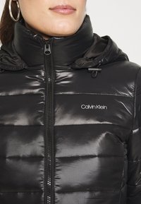 Chaqueta de plumas negra con un acabado brillante, cierre frontal de cremallera, cuello alto y logo de Calvin Klein en el pecho. Cuenta con reguladores en el capó.