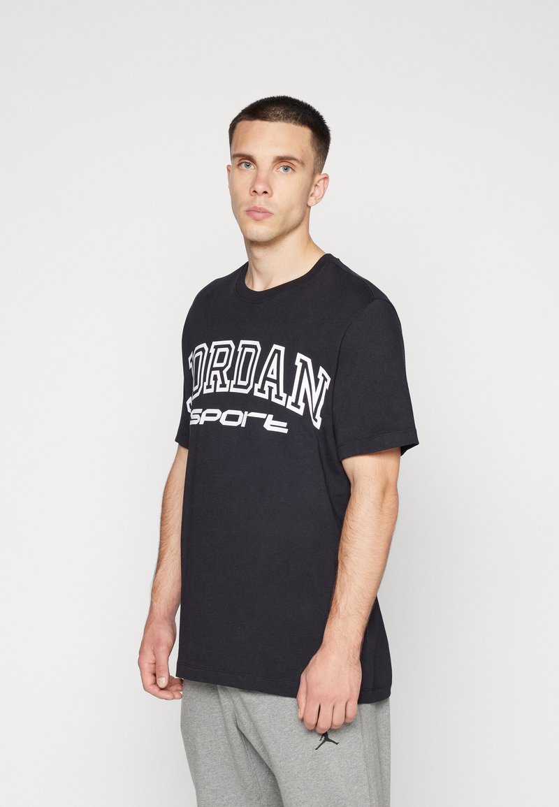 Camiseta de algodón negra con letras blancas "JORDAN Sport", cuello redondo y mangas cortas, combinada con pantalones jogger grises.
