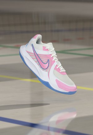 Бяла и розова спортна обувка със синя подметка и лого на Nike, плаваща над пода на фитнес зала с видими цветни линии на игралното поле.