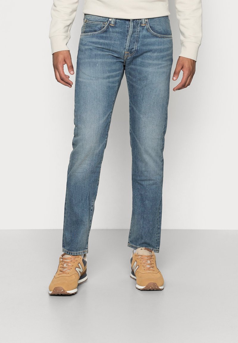 Jeans de mezclilla azul claro con corte recto, que cuentan con cinco bolsillos, un ligero efecto de desgaste y detalles de costura. Llevados con zapatillas color beige.