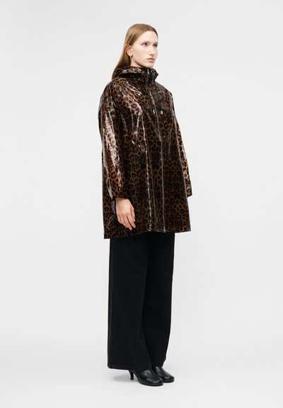 Γυναίκα που φοράει γυαλιστερό αδιάβροχο με κουκούλα σε λεοπαρ σχέδιο, μαύρο παντελόνι με φαρδιά πόδια και μαύρα μποτάκια μέχρι τον αστράγαλο, στέκεται μπροστά σε απλό φωτεινό φόντο.