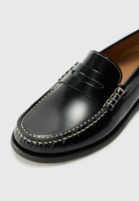 Schwarze Lederloafers mit einer glatten Oberfläche, die über dekorative Nähte und ein erhöhtes Akzentband über dem Spann verfügen. Abgerundete Zehenform.
