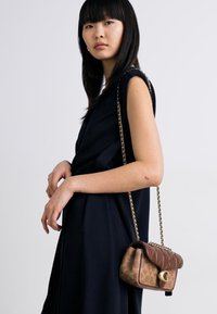 Brown leather handbag with quilted texture and gold hardware, featuring a chain strap. Model wears a black garment, stands against a light background.