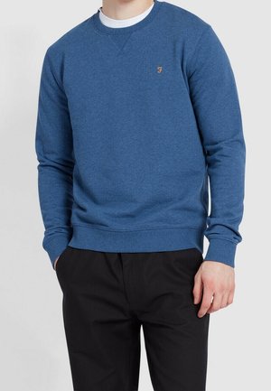 Hombre vistiendo una sudadera azul con cuello redondo y un pequeño logotipo bordado en el pecho, pantalones negros y camiseta blanca, de pie con las manos parcialmente en los bolsillos.