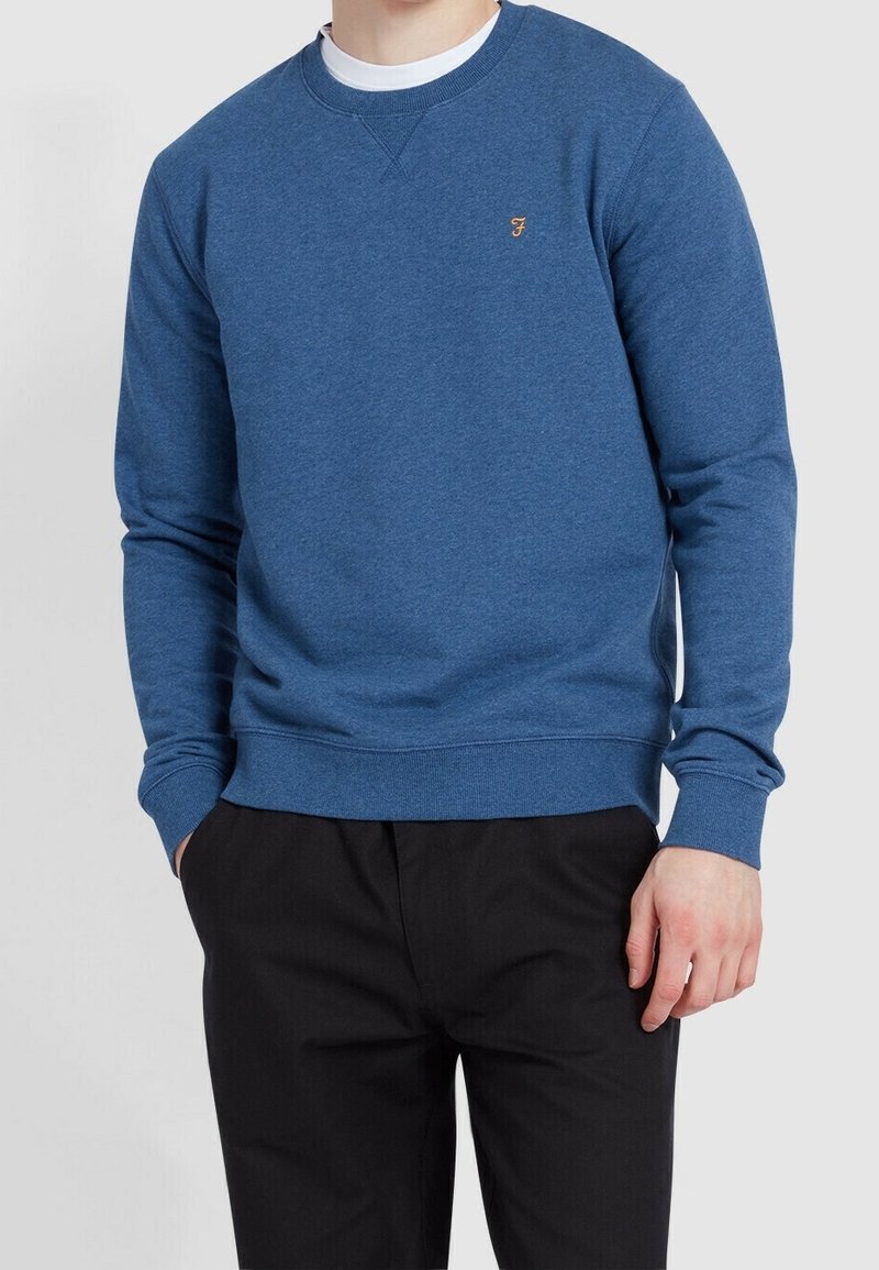Homme portant un sweat-shirt bleu à col rond avec un petit logo brodé sur la poitrine, pantalon noir et sous-vêtement blanc, debout les mains partiellement dans les poches.