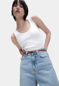 Weißes geripptes Tanktop, kombiniert mit lockeren, hochgeschnittenen hellblauen Jeans. Ein brauner Ledergürtel setzt Akzente, mit glatter Textur und schlichter Gestaltung.