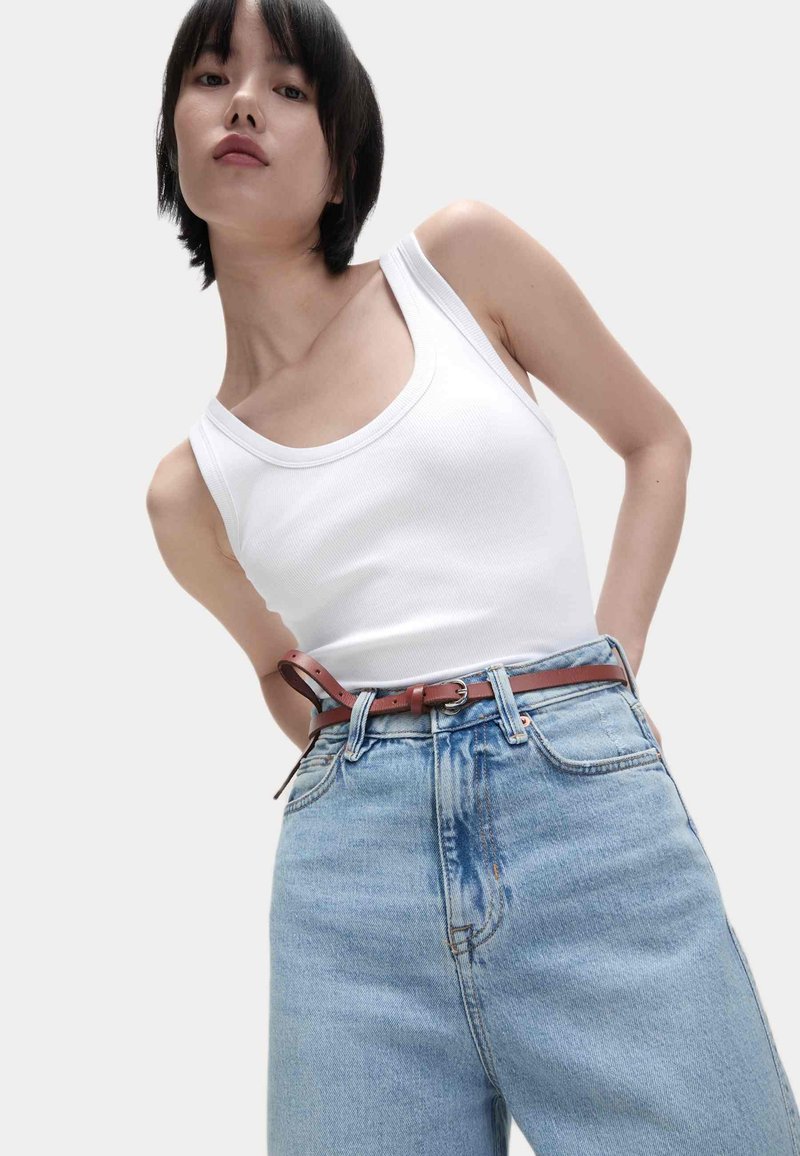 Weißes geripptes Tanktop, kombiniert mit lockeren, hochgeschnittenen hellblauen Jeans. Ein brauner Ledergürtel setzt Akzente, mit glatter Textur und schlichter Gestaltung.