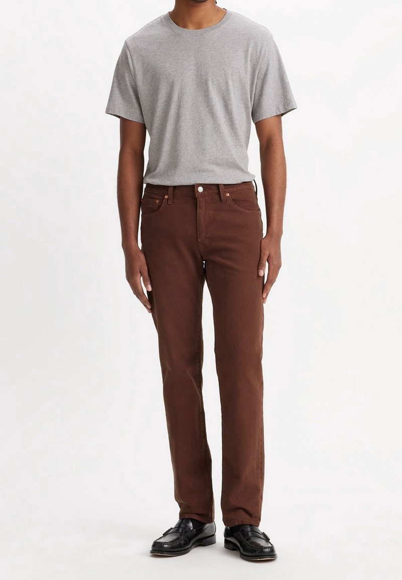 Homme portant un t-shirt gris clair à manches courtes, un pantalon droit marron foncé et des chaussures habillées noires, debout sur un fond blanc.