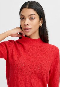 Roter Strickpullover mit hohem Halsausschnitt, der ein Wellenmuster aufweist, kurze Ärmel hat und eine weiche, strukturierte Oberfläche besitzt.