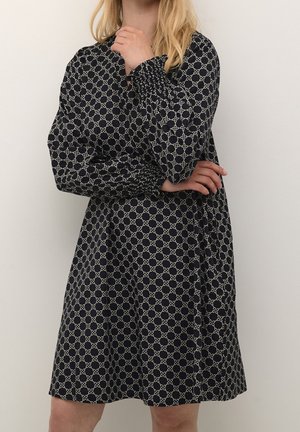 Mujer con un vestido negro hasta la rodilla con un patrón de eslabones blancos, mangas largas abullonadas y puños fruncidos, posando con los brazos cruzados.