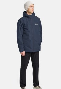 Granatowa wodoodporna kurtka z kapturem, zamkiem z przodu i logo na piersi. Noszona z czarnymi spodniami i kremową czapką beanie.