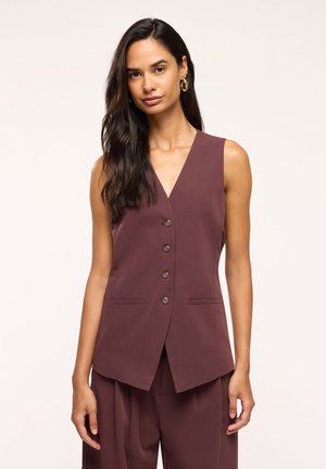Donna con lunghi capelli scuri che indossa orecchini a cerchio dorati e un gilet marrone senza maniche abbottonato con pantaloni marroni abbinati.