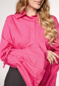 Camicia button-up fucsia con colletto e maniche a campana legate. Il tessuto appare liscio con una trama prominente e un colore vibrante.