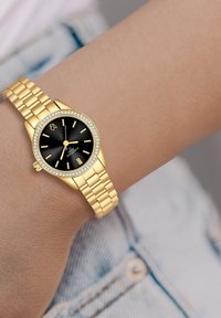 Reloj de pulsera de oro con esfera negra, banda metálica dorada, caja redonda, acentos similares a diamantes y marcadores de hora simples.