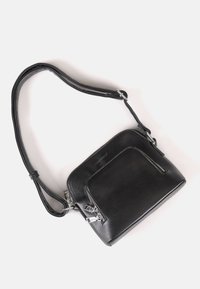 Zwarte leren crossbodytas met verstelbare band, voorvak met rits en zilverkleurige hardware op een witte achtergrond.