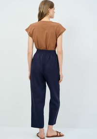 Brun kortärmad topp matchad med marinblå byxor med elastiskt midjeband; båda har en avslappnad passform. Modell med bruna sandaler som står mot en ljus bakgrund.