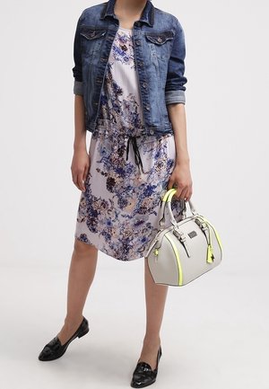 Kvinde iført blomsterkjole med sort snor, blå denimjakke, sorte loafers, der holder en hvid håndtaske med neon gule accenter.