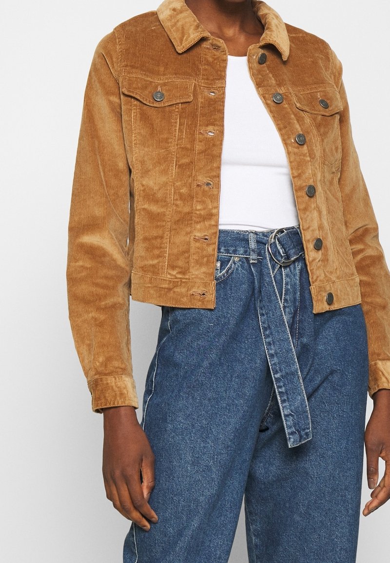 Woman wearing a cropped brown corduroy jacket over a white top and high-waisted blue denim jeans with a fabric belt.