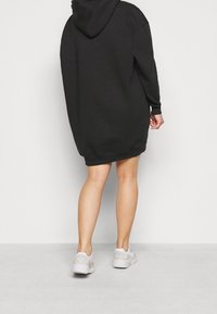 Čern�é oversized mikinové šaty s kapucí, s jemnou texturou a ribbenovými manžetami. Nosené s světle šedými teniskami.