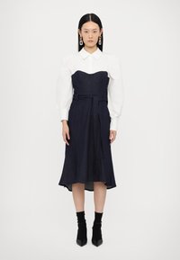 Navy denim jurk met een aansluitende top en een uitlopende rok, met daaronder een witte blouse, een riem in de taille en hoge zwarte enkellaarsjes.