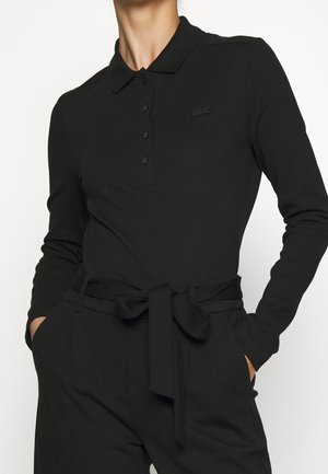 Mujer con camisa polo negra de manga larga metida dentro de pantalones negros de talle alto con un cinturón de tela atado, con las manos en los bolsillos.