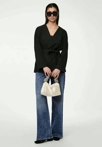Μαύρη μπλούζα τύπου wrap με μακριά μανίκια, συνδυασμένη με τζιν παντελόνι σε φάρδος. Το μοντέλο κρατά μια λευκή τσάντα χειρός με υφή στους χειρολαβές.